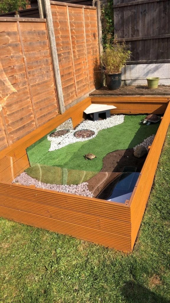 Minimalist sun-deck tortoise garden with fake turf, gravel, and dirt zones against a warm wooden fence